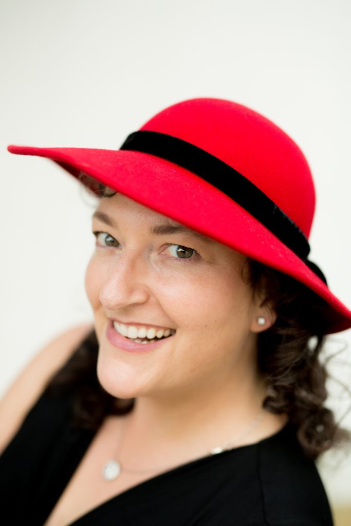 Yvette Keller with a cheeky grin sidways toward the camera, wearing a red felt 1940's hat with a black hatband and a black shirt by Rewind Photography 